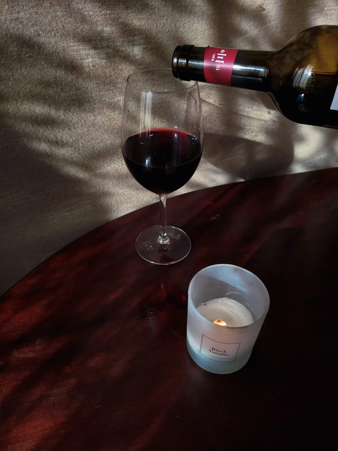 wine glass and a candle are on a table. the wine bottle is over the glass of wine because it finished pouring